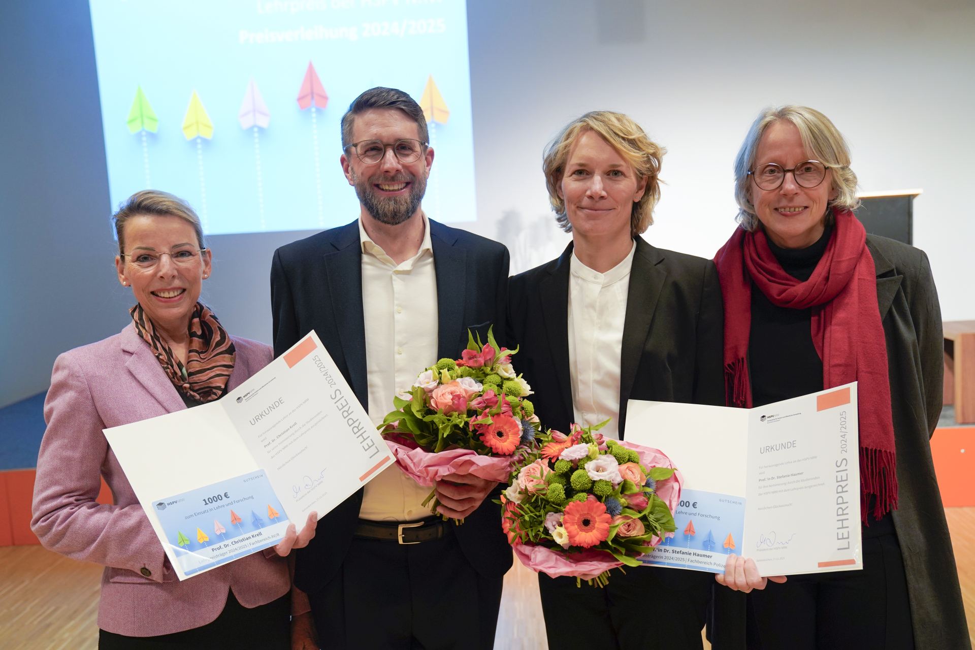 Lehrpreis 2025 Iris Wiesner und Maria Flück stehen links und rechts am Rand und halten die offenen Urkunden hoch. Christian Krell und Stefanie Haumer stehen jeweils mit einem Blumenstrauß zwischen ihnen.