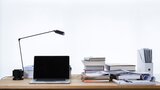 Ein Schreibtisch mit einem Laptop, einer Lampe und einem Stampel Bücher.