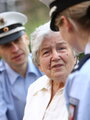 Demenz Eine ältere Frau blickt in die Kamera, links von ihr sieht man das Gesicht eines Polizisten, rechts von ihr eine Polizistin von hinten.